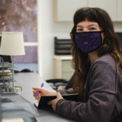 Gaeble Jones, a white woman with brown hair and short bangs, looks up from writing in a notebook while sitting at her desk in the Associated Students of Westminster office