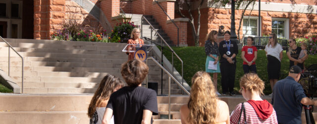President Beth Dobkin stands behind a lectern on the stairs in front of Converse Hall while contributors to this partnership stand beside her to the right.