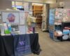 Wide-shot of the Purple Basket shows a table with housing resource information in the form of pamphlets and fliers. Semi-empty shelves line the back wall and a refrigerator stands to the right side against the wall.